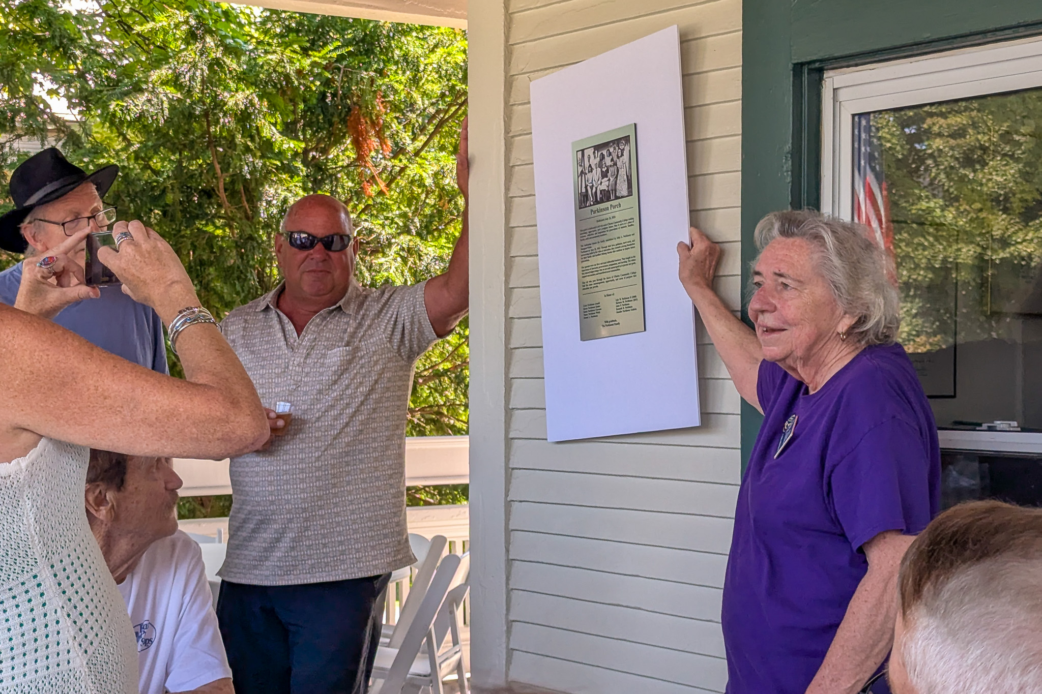 Two people hold up a white board with a plaque on it in front of others on a porch