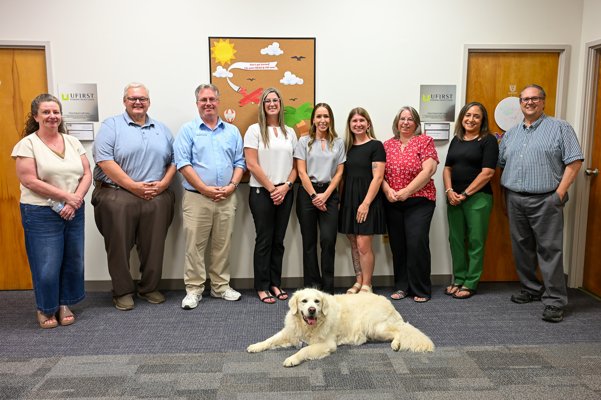 Nine people standing in front of a wall with a dog laying down in front of them